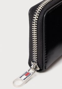 Zbliżenie na czarny róg portfela z srebrnym zamkiem błyskawicznym i taśmą z logo Tommy Jeans w kolorach czerwonym, białym i niebieskim.