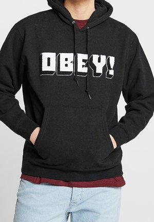 Osoba ubrana w czarną bluzę z kapturem z białymi literami "OBEY!" oraz jasnoniebieskie dżinsy, ręce w przedniej kieszonce.