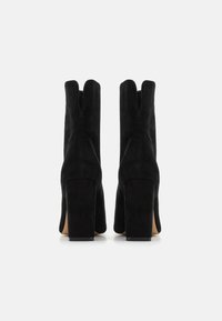 Juodos suede aukštakulnės botai su kvadratine nosimi ir perskirtimu viršuje. Stori blokiniai kulniukai užtikrina stabilumą ir elegantišką siluetą.