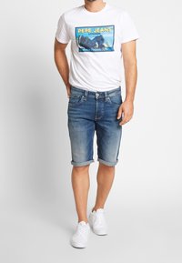 Vit t-shirt med ett blått grafiskt tryck, parat med blå denimshorts med uppvikta fållar, buren med vita sneakers.