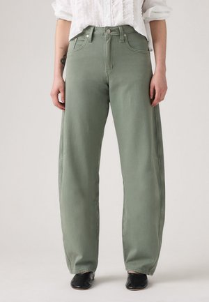 Personne portant un pantalon olive large et taille haute, une chemise blanche à manches longues retroussées et des chaussures noires.