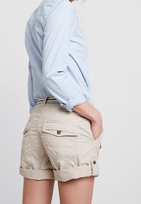 Vaaleansininen kauluspaita yhdistettynä vaaleanbeigeihin cargo-shortsseihin. Shortsseissa on käännetyt lahkeet, taskut ja kultaiset napit. Pehmeä, teksturoitu kangas.
