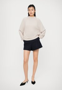 Maglione oversize beige con maniche a raglan, abbinato a pantaloni corti blu navy. Il modello indossa mule nere con punta aperta, in piedi su uno sfondo bianco.