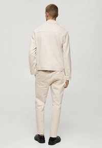 Mand står med ryggen til, iført beige jakke og bukser samt sorte sko foran en ensfarvet grå baggrund.
