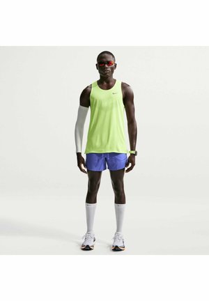 Athlète masculin portant un débardeur vert néon, un short bleu, une manche de compression blanche, des longues chaussettes blanches, des chaussures de course, des lunettes de soleil rouges et une montre de sport.