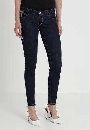 Vaqueros ajustados azul oscuro usados por una persona con zapatos slingback blancos de tacón alto, de pie contra un fondo liso.
