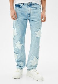 Jeans de un azul claro con grandes parches en forma de estrella y manchas de pintura blanca, usados con zapatillas blancas, enmarcados de la cintura a los pies.