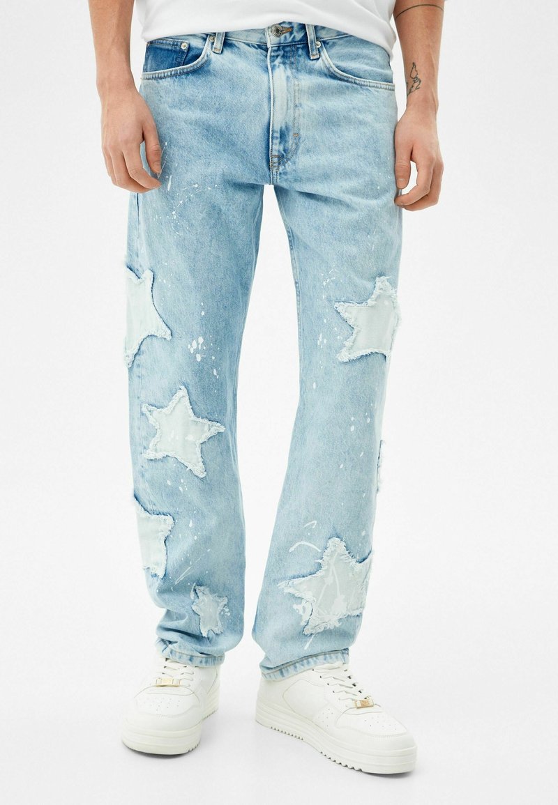 Jeans de un azul claro con grandes parches en forma de estrella y manchas de pintura blanca, usados con zapatillas blancas, enmarcados de la cintura a los pies.