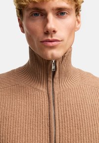 Tan-ribbad stickad zip-up tröja med hög krage och silverdragkedjedetalj. Strukturerad tyg ger ett mysigt utseende.