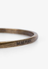 Pulsera de bronce con un diseño suave y redondeado, y un acabado texturizado. Grabada con el texto "SCALPERS" a lo largo de un lado.