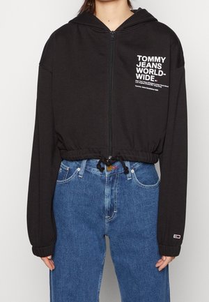Zwarte cropped zip-up hoodie met witte tekst "Tommy Jeans Worldwide" op de borst, gecombineerd met hoog getailleerde blauwe spijkerbroek.