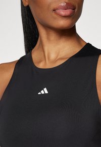 Top sportiv de culoare neagră, confecționat dintr-un material neted și elastic. Prezintă un logo alb și bretele subțiri, conceput pentru confort și libertate de mișcare.
