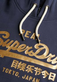 Marine hoodie met gouden tekst "SuperDry" en Japanse karakters, met witte trekkoordjes met gemarkeerde metalen tips.