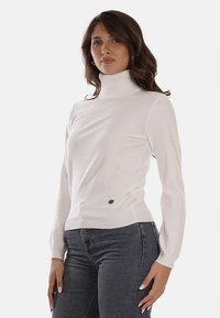Maglione bianco a collo alto con maniche lunghe, caratterizzato da una consistenza morbida e sottili brillanti. Accentuato da un piccolo bottone decorativo sul lato.