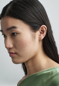 Pendiente de oro en forma de espiral, llevado en la oreja izquierda. La modelo tiene el cabello largo y oscuro y lleva una blusa verde de satén.