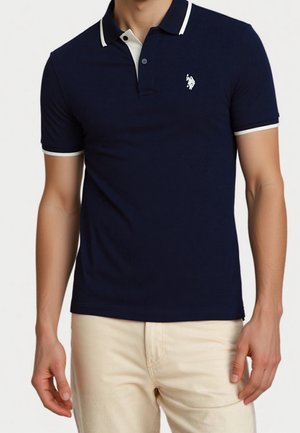 Mand iført navyblå poloshirt med hvide kanter og beige bukser, vist fra nakken til midt på låret mod en ensfarvet baggrund.