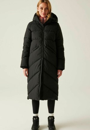 Kobieta w długim czarnym pikowanym zimowym płaszczu z kapturem, czarnych legginsach, białych skarpetkach i czarnych butach trekkingowych stoi na jednolitym tle.