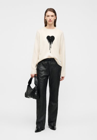 Νεαρή γυναίκα που φοράει ένα κρεμ πουλόβερ με ένα μαύρο καρδιά που στάζει γραφικό σχέδιο, μαύρο δερμάτινο παντελόνι, μαύρα μποτάκια και κρατά μια μαύρη τσάντα.