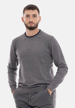 Uomo con capelli corti e scuri e barba, indossa un maglione grigio a maniche lunghe e pantaloni scuri, in piedi con le mani nelle tasche.