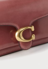 Καφέ δερμάτινη τσάντα χειρός με ομαλή υφή, που διαθέτει εξέχον κούμπωμα σε χρυσή απόχρωση και αντίθετη ραφή κατά μήκος των άκρων.