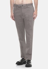 Uomo che indossa pantaloni grigi a taglio dritto e mocassini in pelle nera, in piedi davanti a uno sfondo bianco semplice.