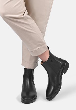 Person wearing black leather ankle boots with elastic side panels and beige rolled-up pants on a plain background.