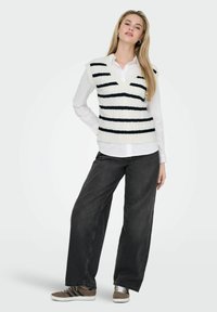 White knit vest with navy stripes over a white collared shirt, paired with loose black jeans and brown sneakers featuring black stripes.