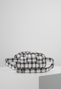 Sac à carreaux noir et blanc en matériau synthétique, forme rectangulaire, bandoulière réglable et accents métalliques argentés.