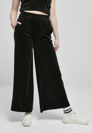 Svarte, vide bukser laget av myk velur, med elastisk midje og sidelommer. Brukt med hvite joggesko og stripete sokker.