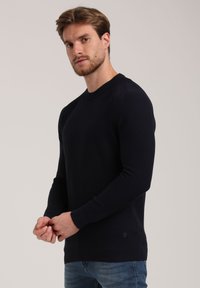 Dunkelblauer Pullover aus strukturiertem Stoff mit gerippten Bündchen. Verfügt über einen Rundhalsausschnitt und einen schlanken Schnitt, kombiniert mit blauen Jeans.