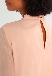 Blouse rose en tissu texturé avec un col à volants et une fermeture boutonnée à l'arrière, présentant un design en forme d'œillet.