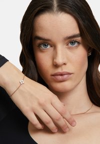 Bracciale in oro rosa con una delicata catena, caratterizzato da tre piccole perle bianche raggruppate insieme. La mano della modella è posata sulla clavicola.