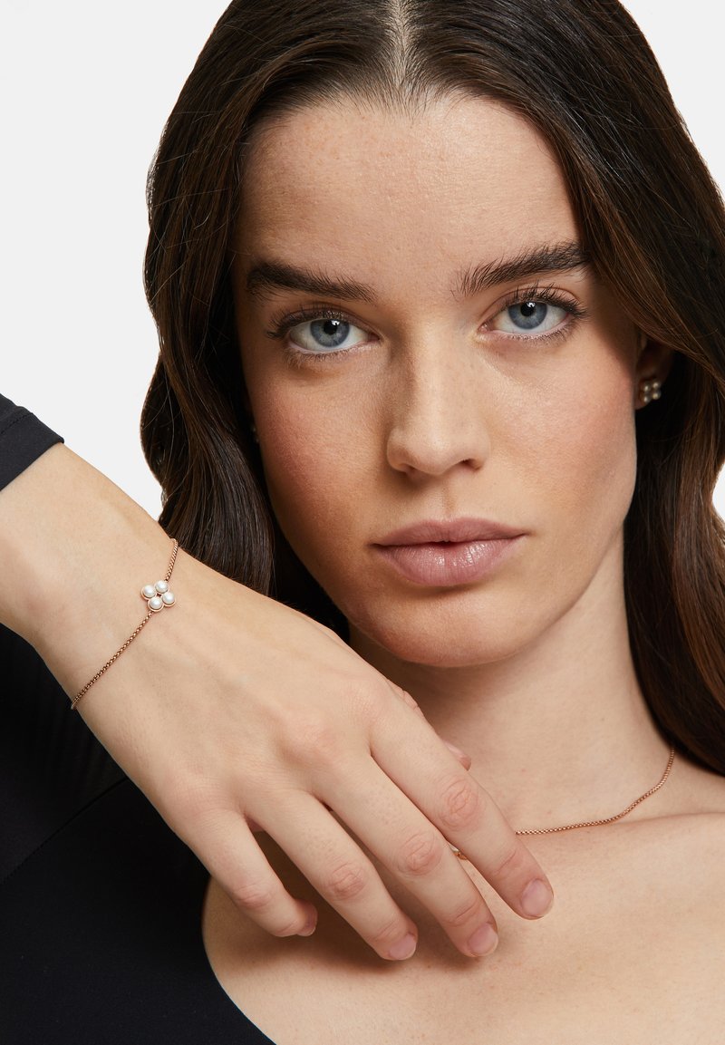 Bracciale in oro rosa con una delicata catena, caratterizzato da tre piccole perle bianche raggruppate insieme. La mano della modella è posata sulla clavicola.