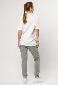 Ponytailiga naine, kes kannab valget lahtist T-särki, halli dressipükse, valgeid tenniseid, kuldseid rõngakujulisi kõrvarõngaid ja käevõru, seistes seljaga vaataja poole.