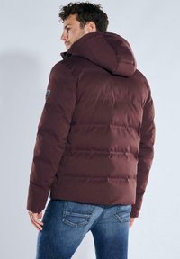Bordeauxrote gesteppte Jacke mit Kapuze, die horizontale Steppnähte und ein Logo-Patch am Ärmel aufweist. Wird mit blauer Jeans getragen.
