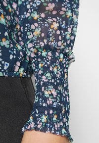 Blusa trasparente blu navy con maniche e polsini arricciati, decorata da un motivo floreale multicolore, indossata sopra un tessuto di denim nero.