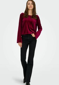 Burgundy sammetstopp med utsvängda ärmar, kombinerat med svarta utsvängda jeans. Tyget har en slät glans och en avslappnad passform.