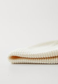 Un bonnet en tricot crème plié avec une texture côtelée. Il est fabriqué dans un matériau doux et confortable, et est arrondi sur le dessus.