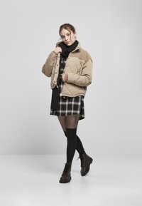 Beige gesteppte Jacke mit strukturiertem Schulterbereich, getragen über einem schwarz-weißen karrierten Kleid, schwarzem Schal und knielangen schwarzen Socken mit braunen Stiefeln.