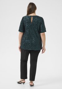 O femeie îmbrăcată într-un top din paiete verde închis, cu un decolteu pe spate, pantaloni negri și pantofi slingback cu toc mic negri, stând cu spatele.