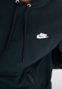 Svart Nike-hoodie med en vit logo, som har en dragkedjeficka och snoddar vid kragen. Mjuk material med en texturerad yta.