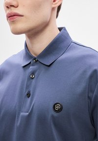 Modré polo tričko s klasickým golierom, tromi čiernymi gombíkmi a okrúhlym čiernym logom na ľavej strane hrudníka. Hladká textúra.