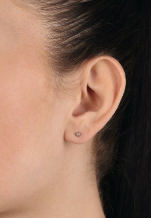 Boucles d'oreilles en argent, petite forme ronde, texture lisse, enfilées dans une oreille percée. Teinte de peau visible, cheveux tirés en arrière. Design minimaliste.