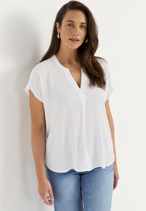 Kvinde med langt brunt hår, iført sølvfarvede øreringe i ringform, en løs hvid kortærmet bluse og lyseblå jeans, stående op ad en hvid væg.