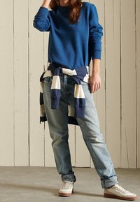 Suéter de punto azul con cuello redondo, vaqueros azul claro y zapatillas blancas con suelas color caqui. Top a rayas azul marino y blanco atado a la cintura.