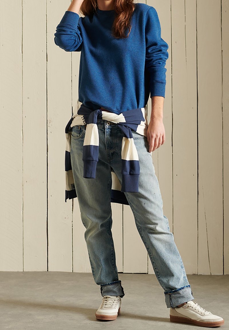 Suéter de punto azul con cuello redondo, vaqueros azul claro y zapatillas blancas con suelas color caqui. Top a rayas azul marino y blanco atado a la cintura.