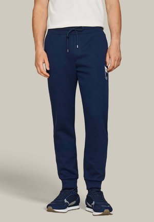 Hombre con pantalones deportivos azul marino con cordón, logo en el muslo, camiseta blanca y zapatillas azul marino, de pie frente a un fondo liso.