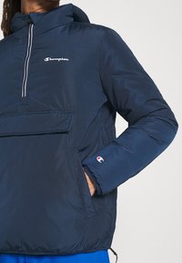 Chaqueta de forro polar de color navy con un bolsillo delantero, cuello alto y logo de Champion. Cuenta con puños elásticos y un dobladillo con cordón para ajuste.
