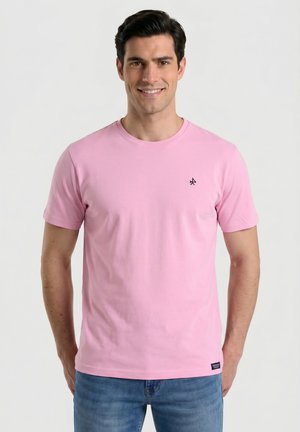 Hombre de pie sonriendo, lleva una camiseta rosa de manga corta con pequeño logo negro y vaqueros azules, fondo claro liso.