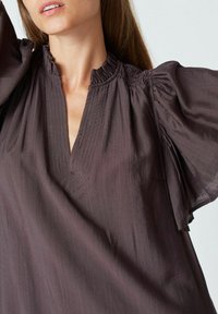 Bluză de culoare prună, cu decolteu în V, guler cu volane și mâneci bufante. Confecționată dintr-un material ușor și neted, cu linii verticale subtile.